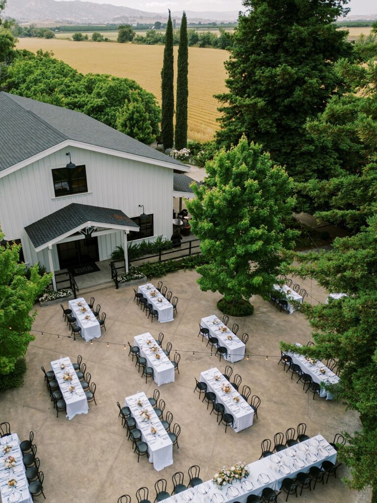 An outdoor wedding setup with rows of tables and chairs arranged on a patio near a white building, surrounded by trees and fields.