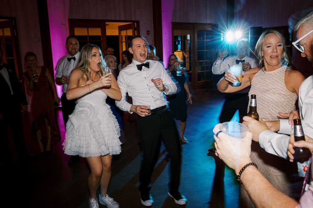 People in formal attire dance and hold drinks at a lively indoor party, with colorful lighting and a bright flash in the background.