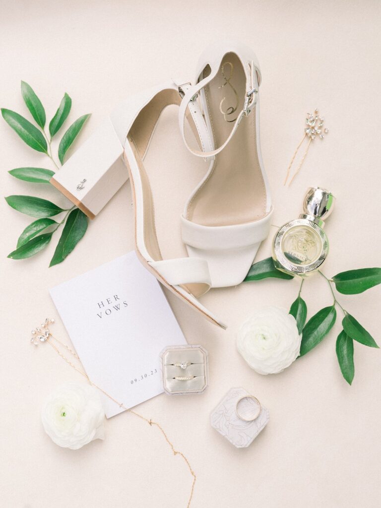 Flat lay of wedding accessories: white high-heeled shoes, perfume, ring, floral decoration, vow booklet with "HER VOWS 09.30.23," and a small box on a light background.