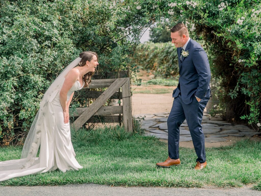 A bride in a white dress and veil laughs while leaning forward. A groom in a blue suit and brown shoes stands facing her, smiling outdoors near greenery and a wooden gate.