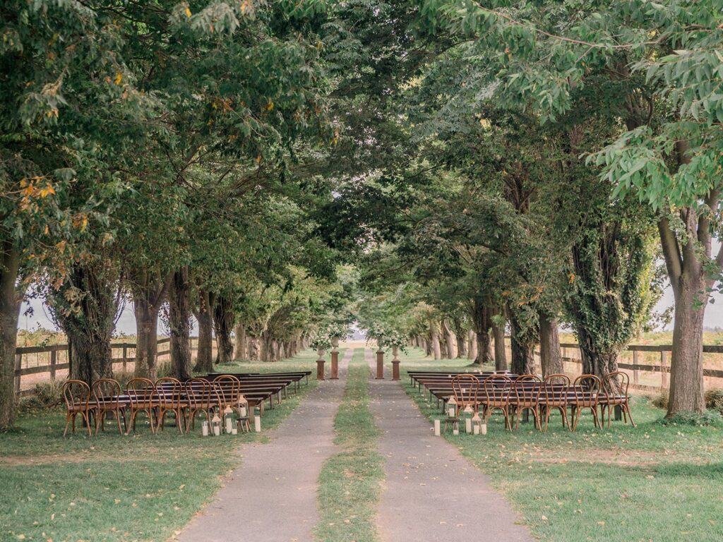 Outdoor wedding ceremony setup with wooden chairs and benches arranged in rows beneath a canopy of trees. Candles in glass holders line the aisle on a path.
