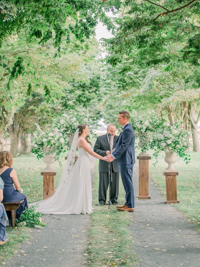 A couple stands at an outdoor wedding ceremony, holding hands in front of an officiant. They are surrounded by greenery and floral arrangements.