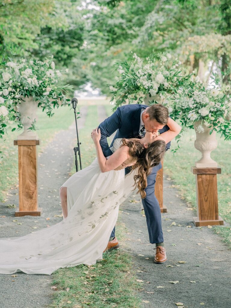 A couple kisses on a tree-lined path, with floral arrangements on pedestals in the background. The bride's dress has a long train.