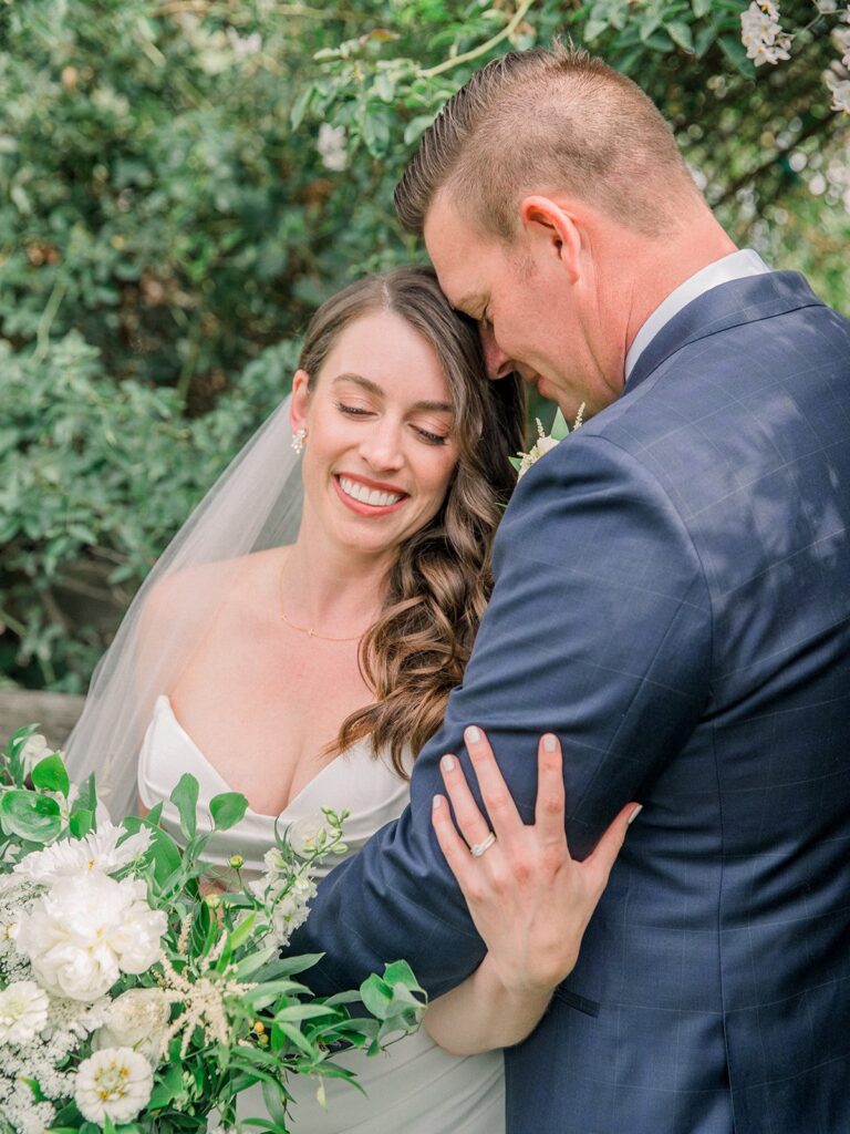 A bride and groom embrace outdoors with greenery and white flowers. The bride smiles with her eyes closed, and the groom wears a dark suit.