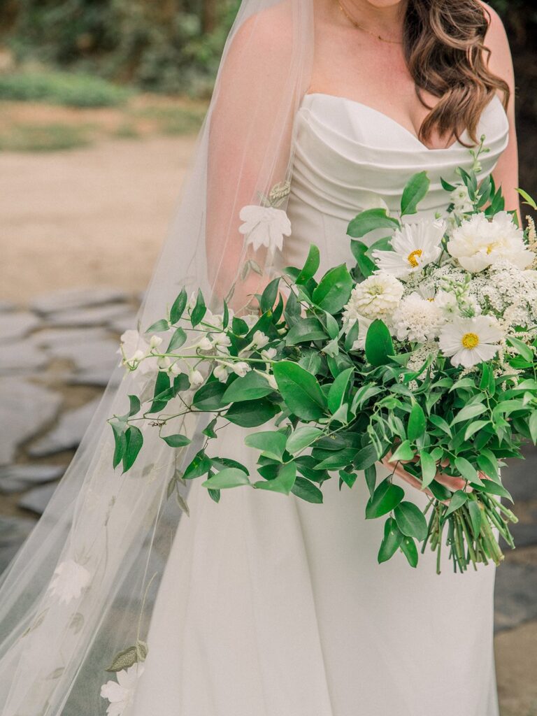 Bride holding a bouquet of white flowers and green leaves, wearing a white strapless gown with a long veil adorned with flowers. Background features stone path and greenery.