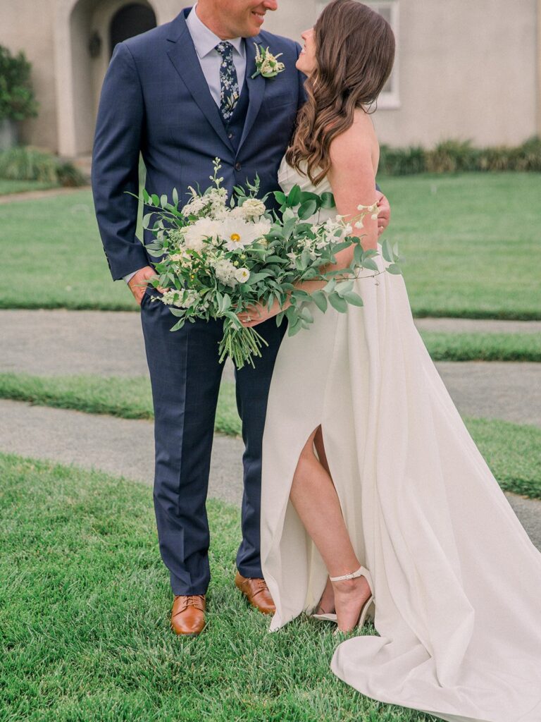 Bride and groom stand on grass, holding a bouquet of flowers. Bride wears a white dress with a slit, groom in a navy suit. House in the background.