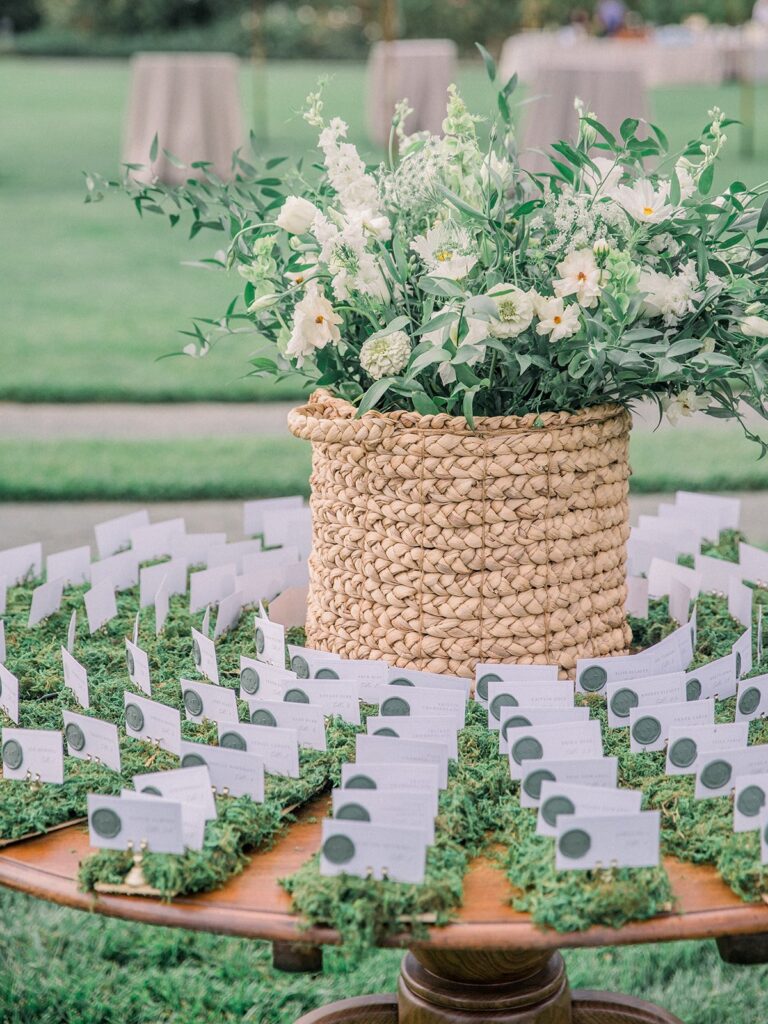 A round table displays a woven basket with white flowers and greenery, surrounded by name cards arranged on a moss-covered surface, set outdoors on a grassy lawn.
