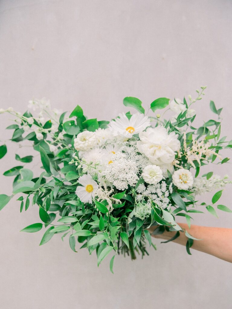 A hand holding a bouquet of white flowers and greenery against a plain background.