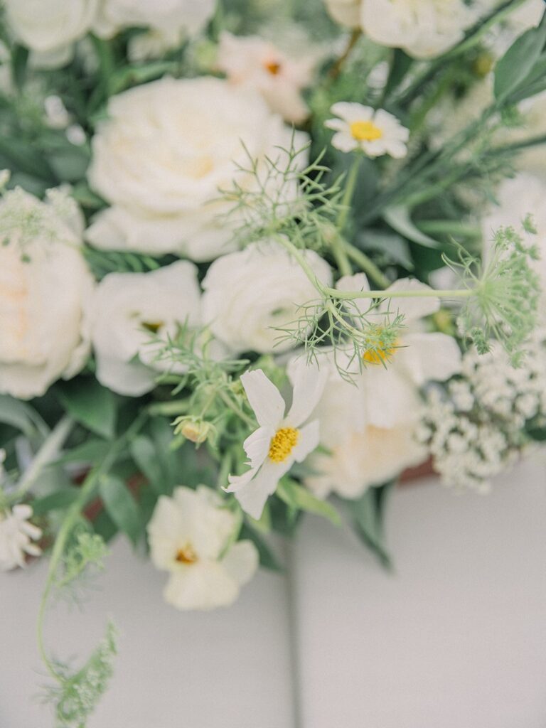 A close-up of a floral arrangement featuring white roses and delicate white daisies with yellow centers, surrounded by green foliage on a light background.