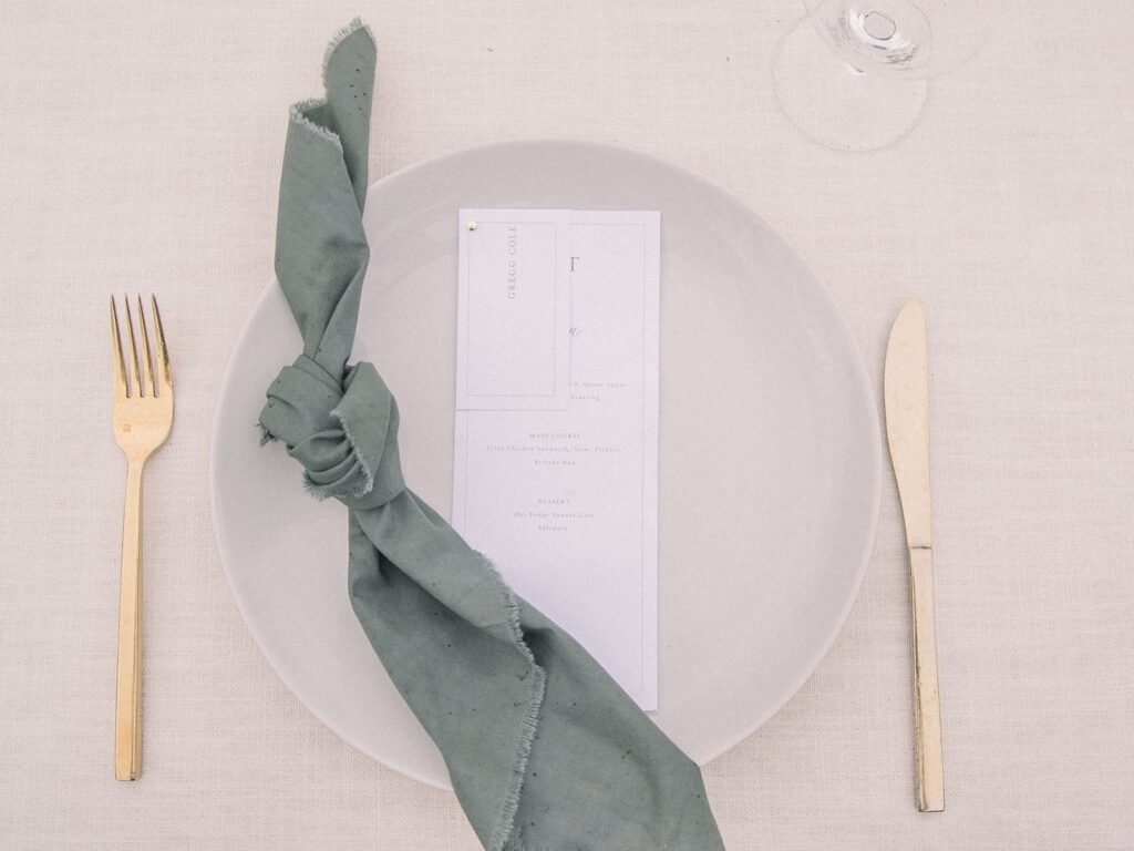 A white plate with a tied green napkin and a menu card. A fork and knife are placed on either side.