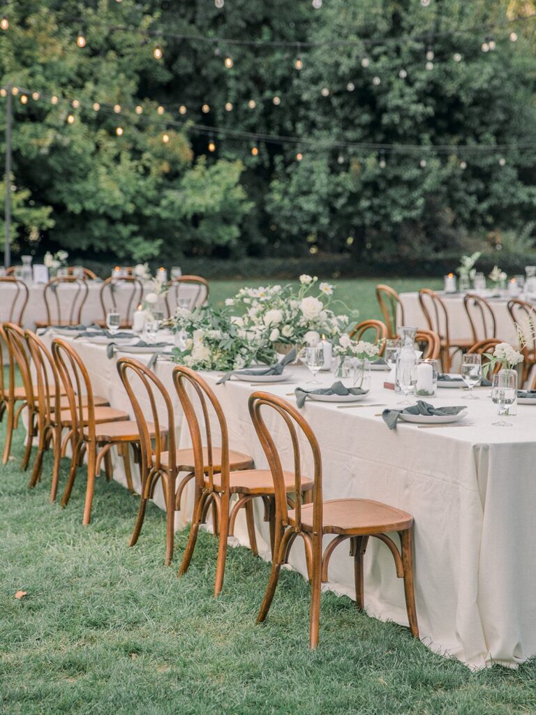 Outdoor wedding reception setup with long tables covered in white tablecloths, wooden chairs, floral centerpieces, and string lights overhead.