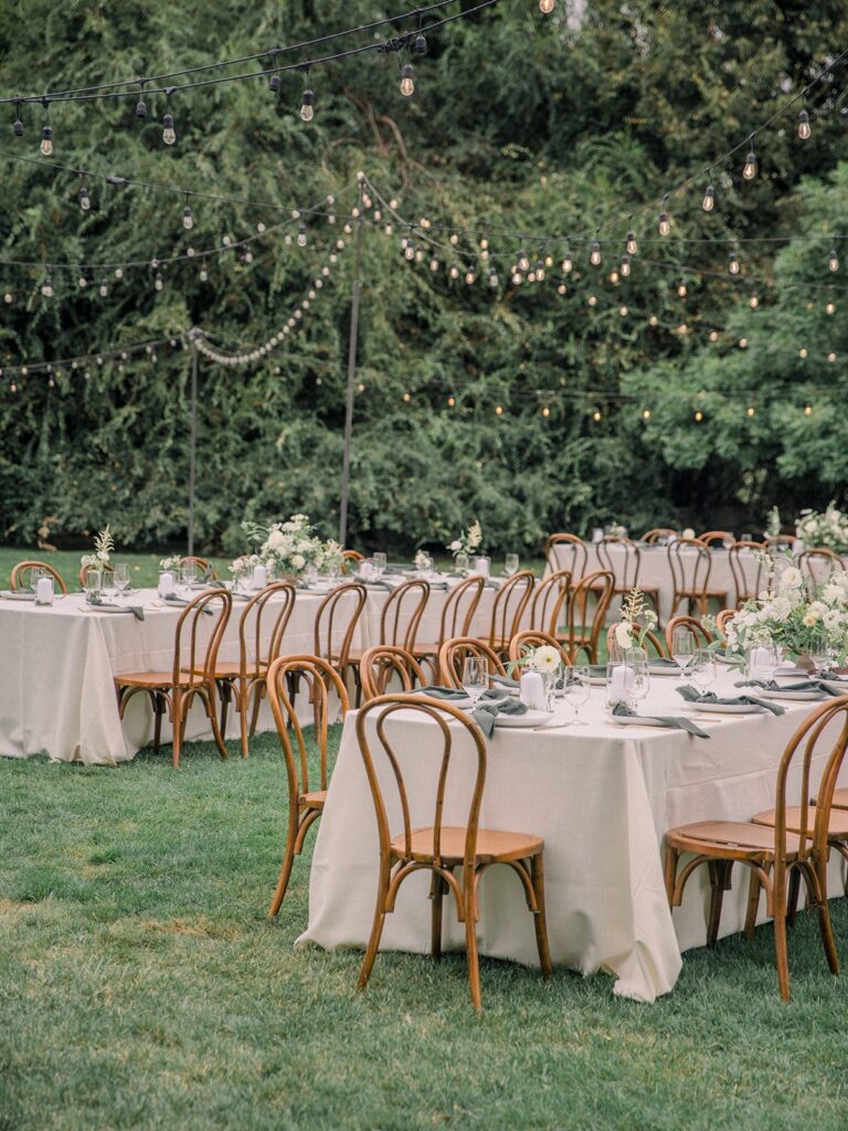 Outdoor event with long tables and wooden chairs set on grass. Tables are covered with white cloths, decorated with flowers. String lights are hung above, surrounded by trees.