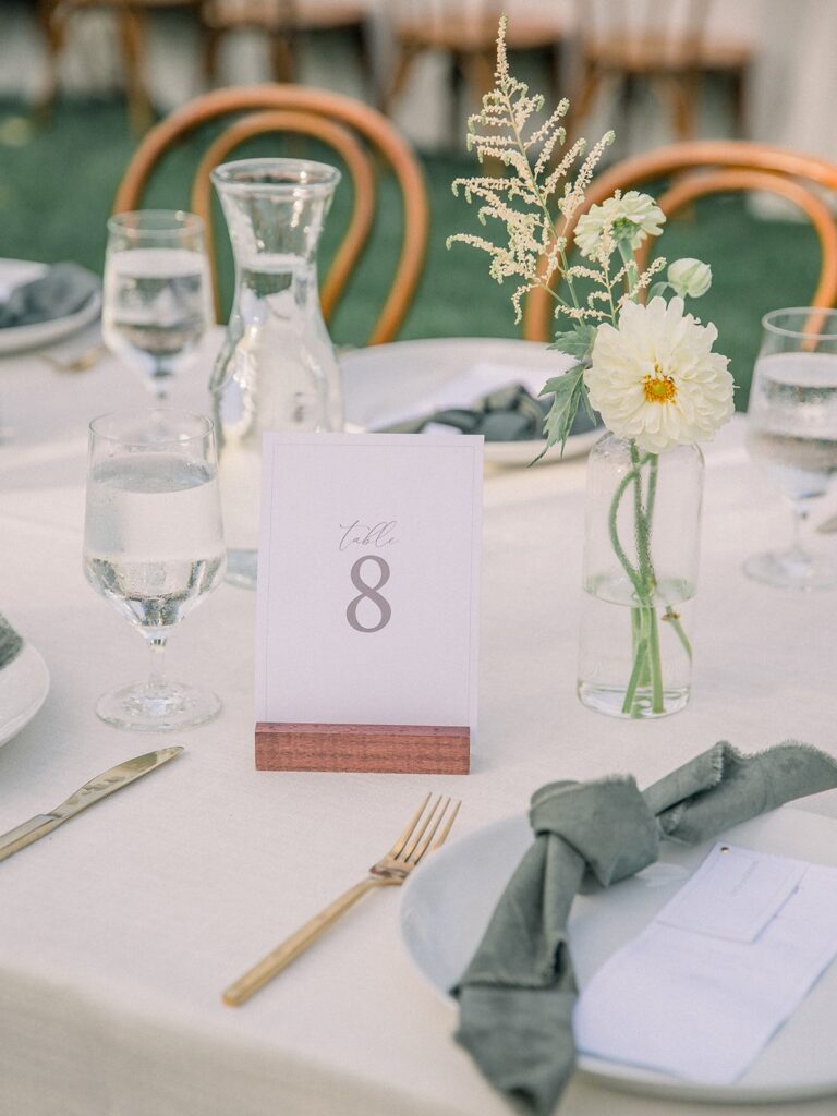 Dining table set for an event with a table number 8 card, glassware, white plates, green napkins, and a small floral arrangement in a glass vase.