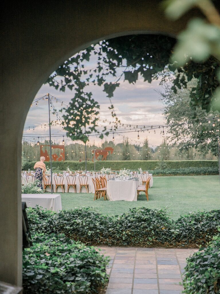 Outdoor event space with decorated tables and chairs under string lights, viewed through an archway with greenery.