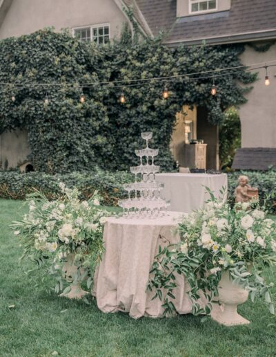 A tiered champagne tower on a table, surrounded by floral arrangements and greenery, set outdoors near a vine-covered house with string lights above.