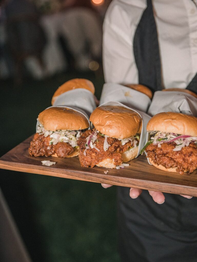 A person holds a wooden tray with multiple fried chicken sandwiches, each topped with coleslaw and wrapped partially in white paper.
