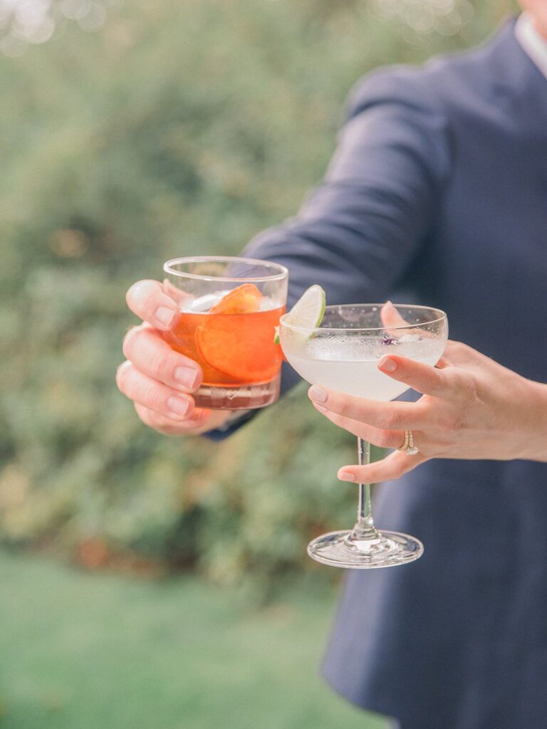 Two hands holding cocktails; one with an orange slice in a tumbler, the other with a lime wedge in a coupe glass. Background is blurred greenery.
