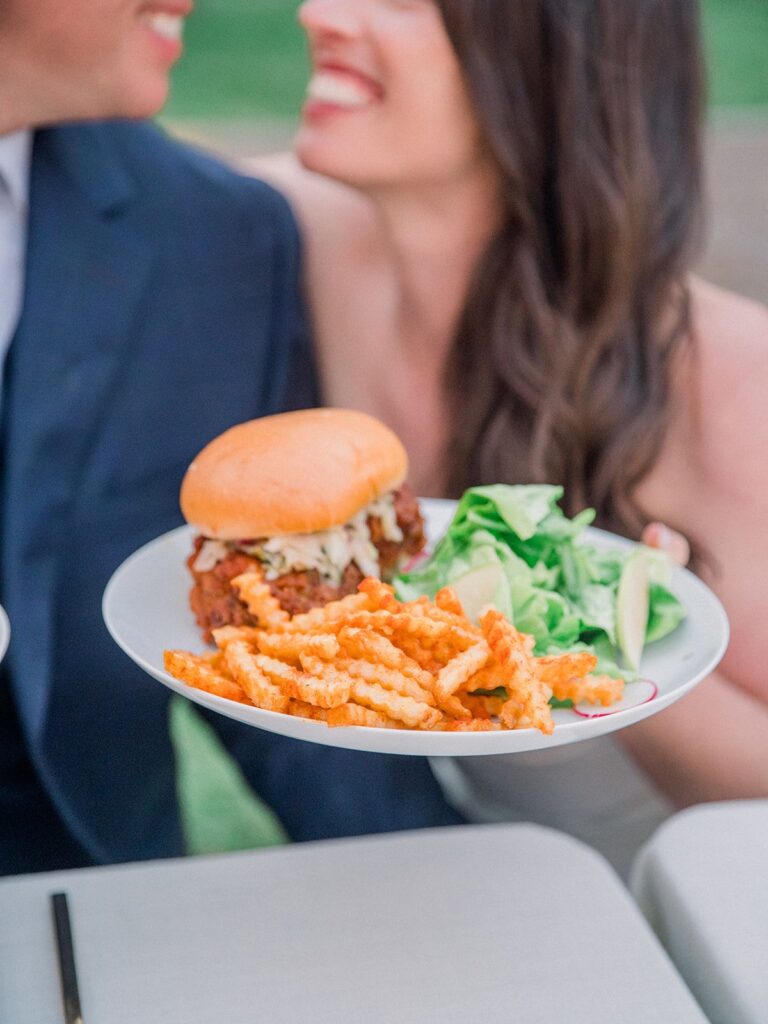 A person holds a plate with a burger, crinkle-cut fries, and a green salad. Two people are smiling in the background.