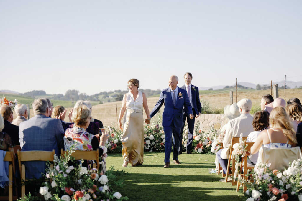 A couple walks down an outdoor aisle, holding hands, surrounded by seated guests and floral arrangements, with a landscape in the background.