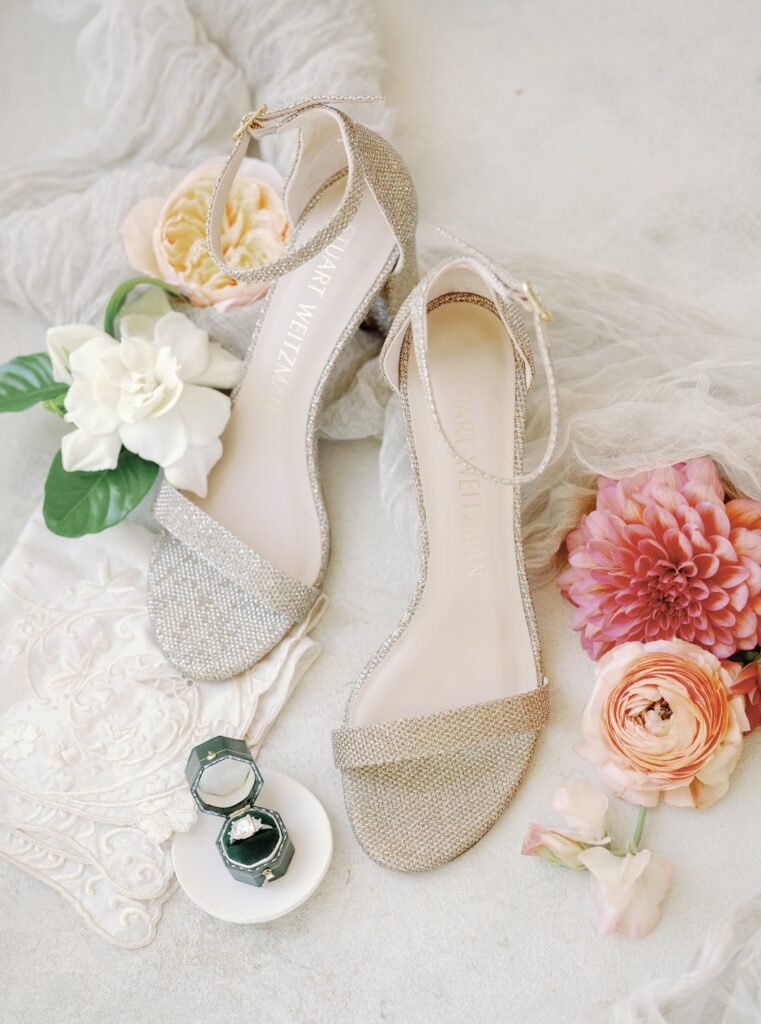 Sparkly high-heeled sandals with floral accents, lace fabric, and two small jewelry boxes on a light background.