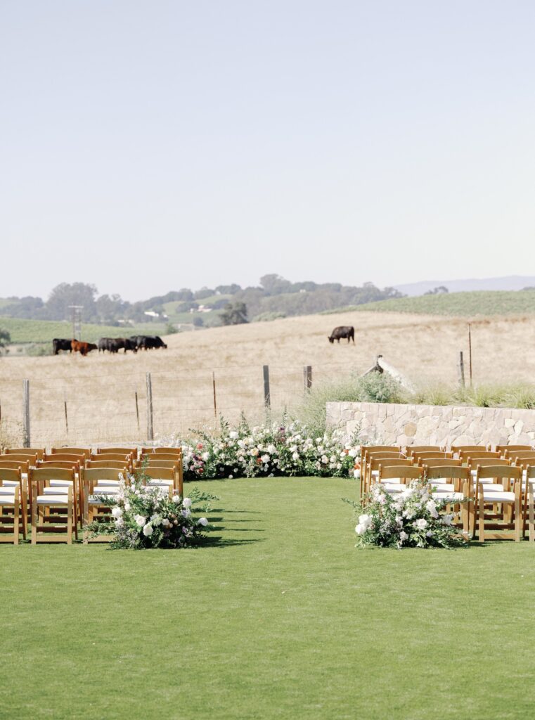 A wedding ceremony setup with wooden chairs and floral arrangements on a green lawn, with cows grazing in a distant field.