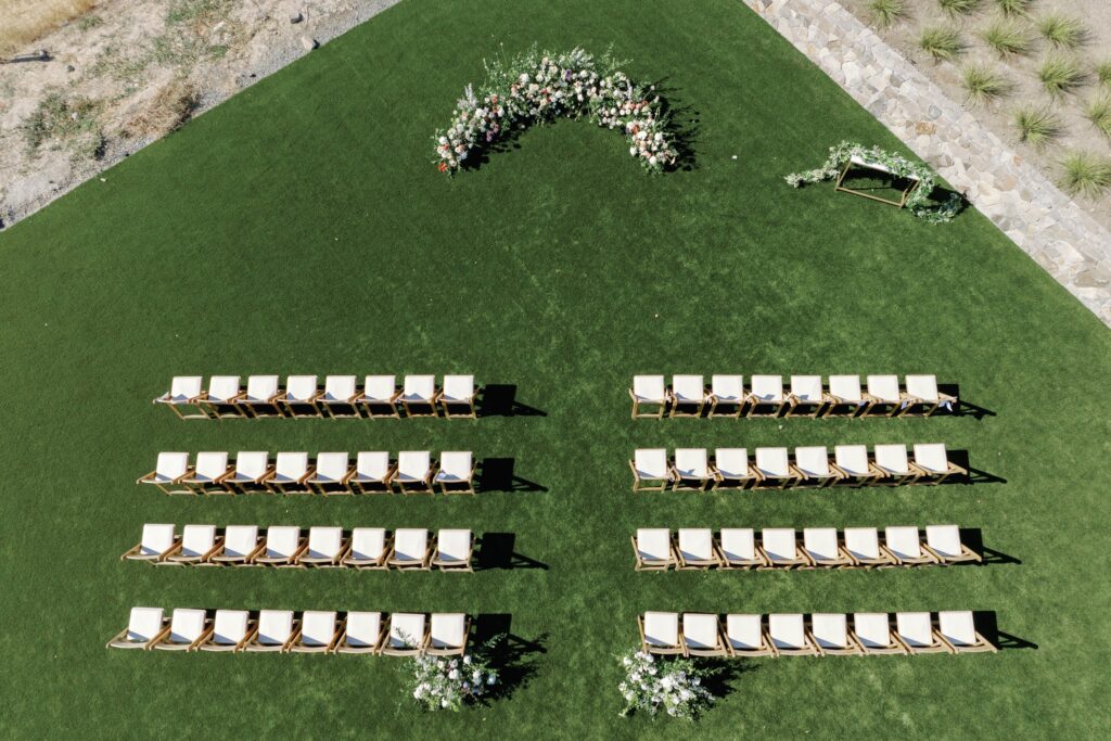 Aerial view of a wedding ceremony setup on a green lawn with rows of white chairs and a floral arch at the front.