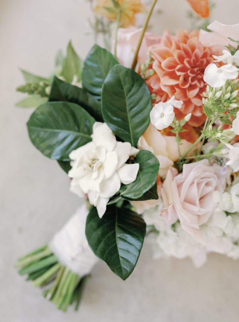 A bouquet with white gardenias, pink roses, orange dahlias, green leaves, and assorted flowers wrapped in white ribbon.
