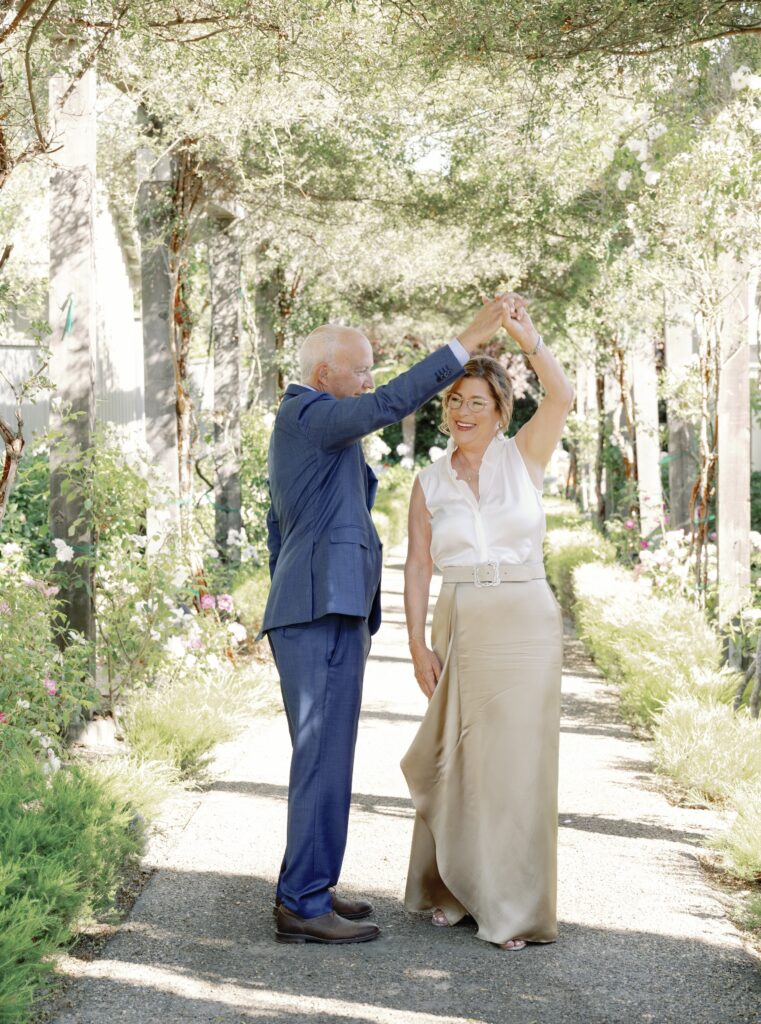 An elderly man in a blue suit and a woman in a white blouse and beige skirt dance together in a garden pathway.