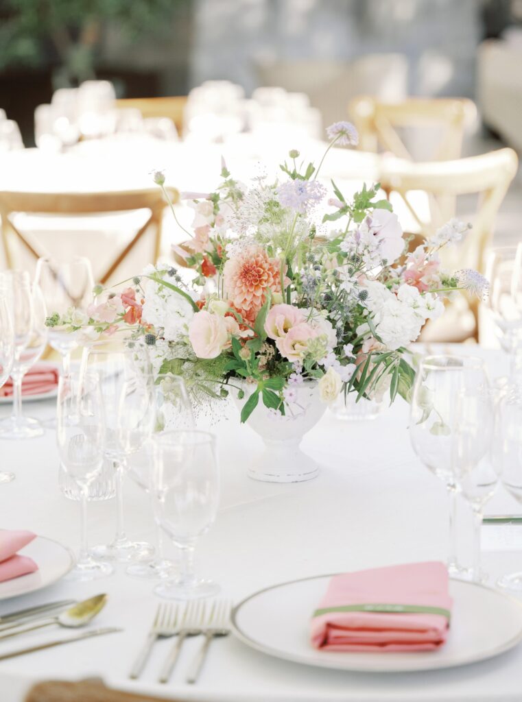 A floral centerpiece with pink and white flowers on a table set with plates, pink napkins, glassware, and cutlery.