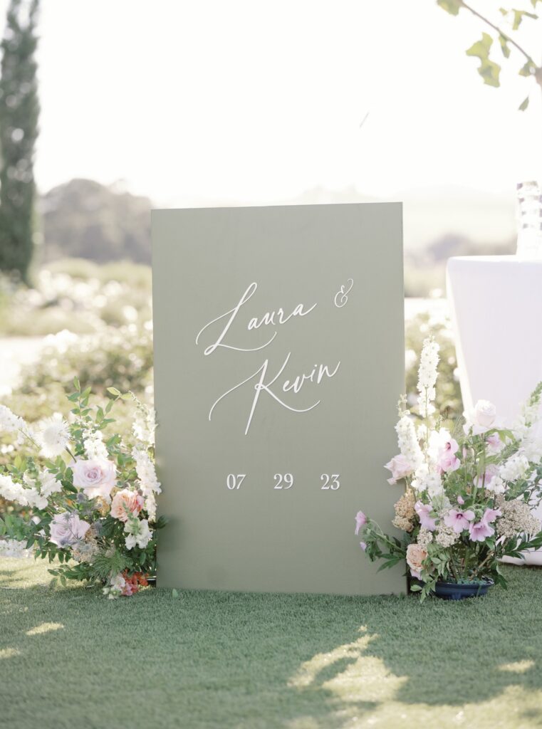 A green wedding sign reads "Laura & Kevin" with the date "07 29 23," surrounded by flowers.