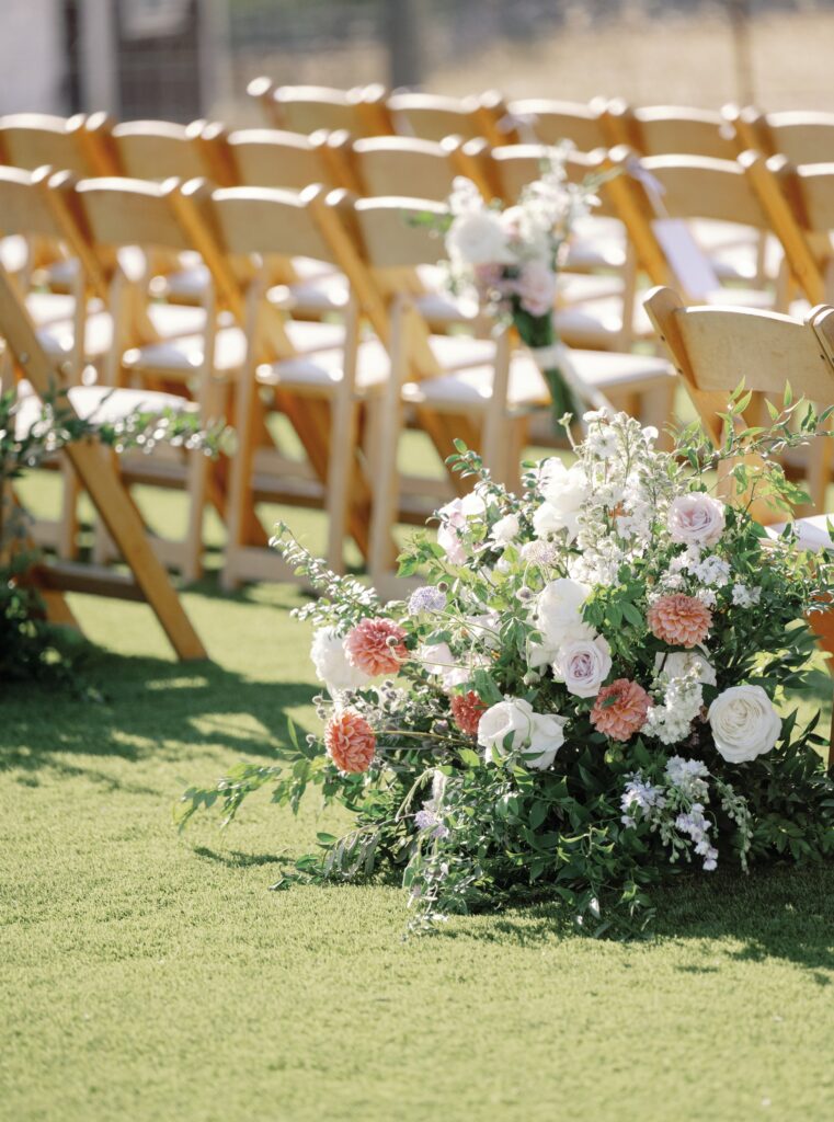 Wooden chairs are arranged in rows on a grassy area. Floral arrangements with white and pink roses decorate the aisle.