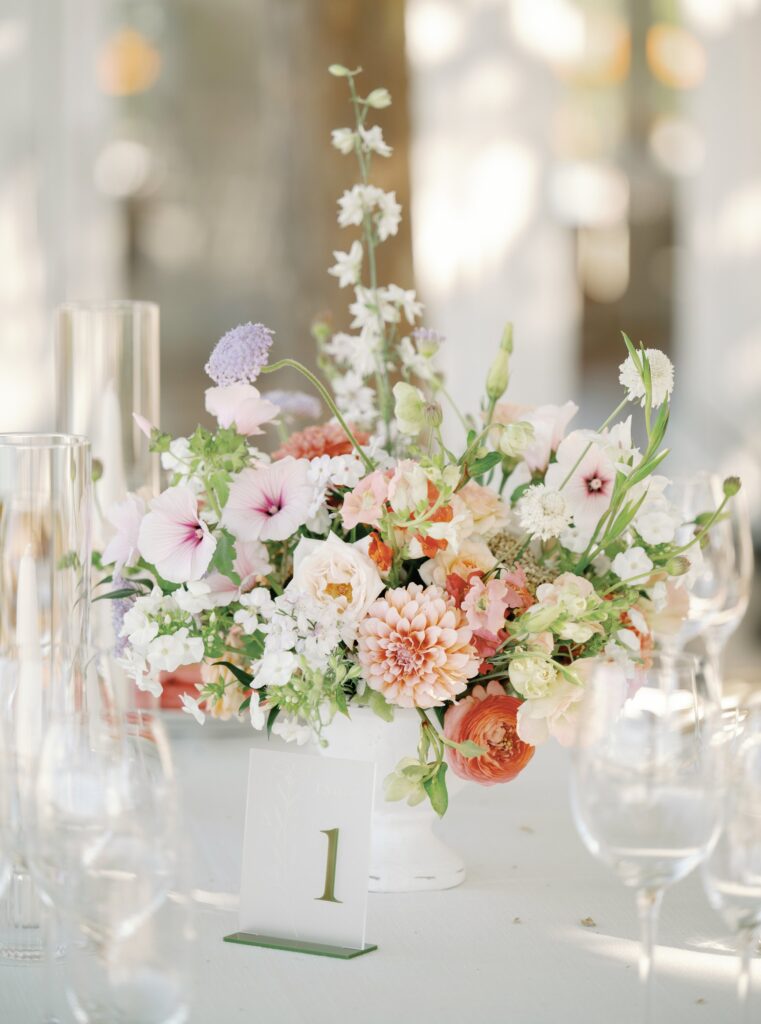 A floral centerpiece with pink, white, and peach flowers in a vase, placed on a table, with clear glassware and a table number card labeled "1".