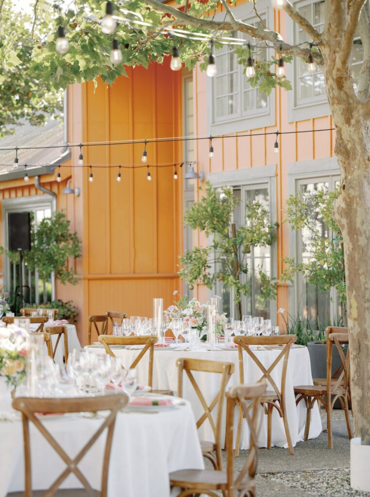 Outdoor dining area with tables set for an event. Round tables with white tablecloths and wooden chairs, under string lights, next to an orange building.