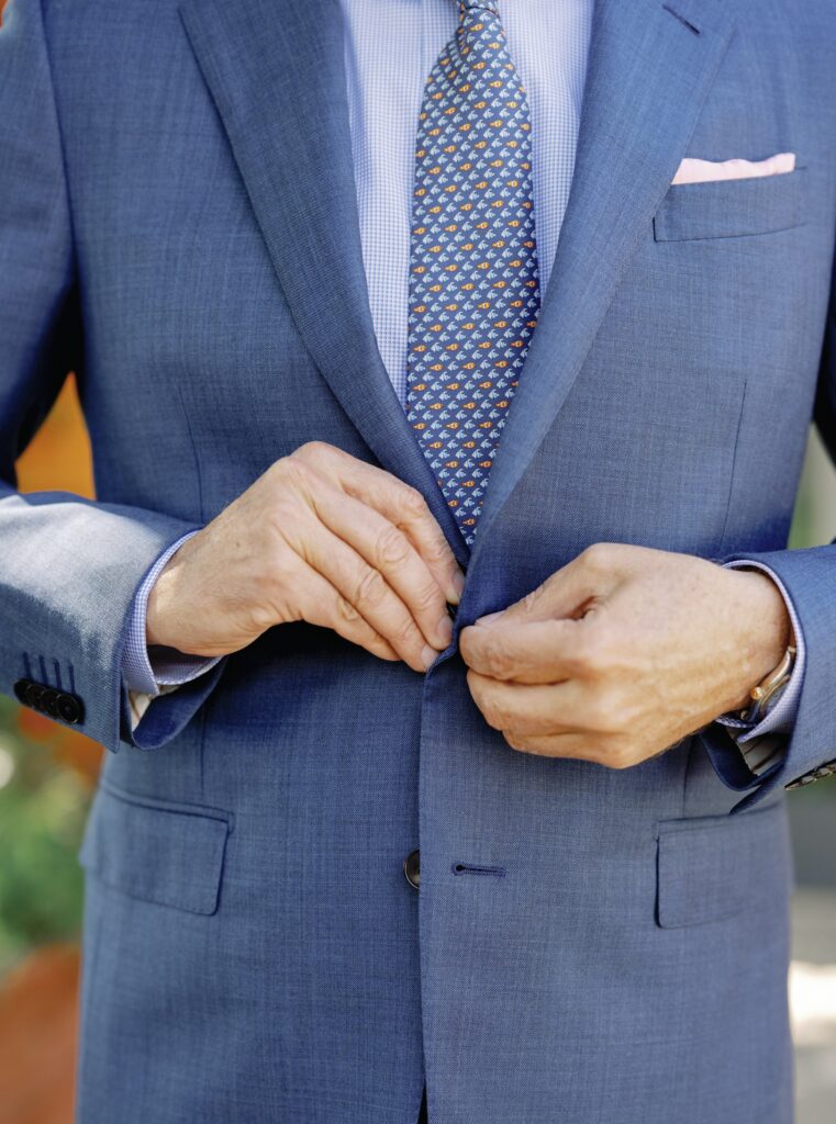 A person wearing a blue suit and patterned tie adjusts their jacket button.