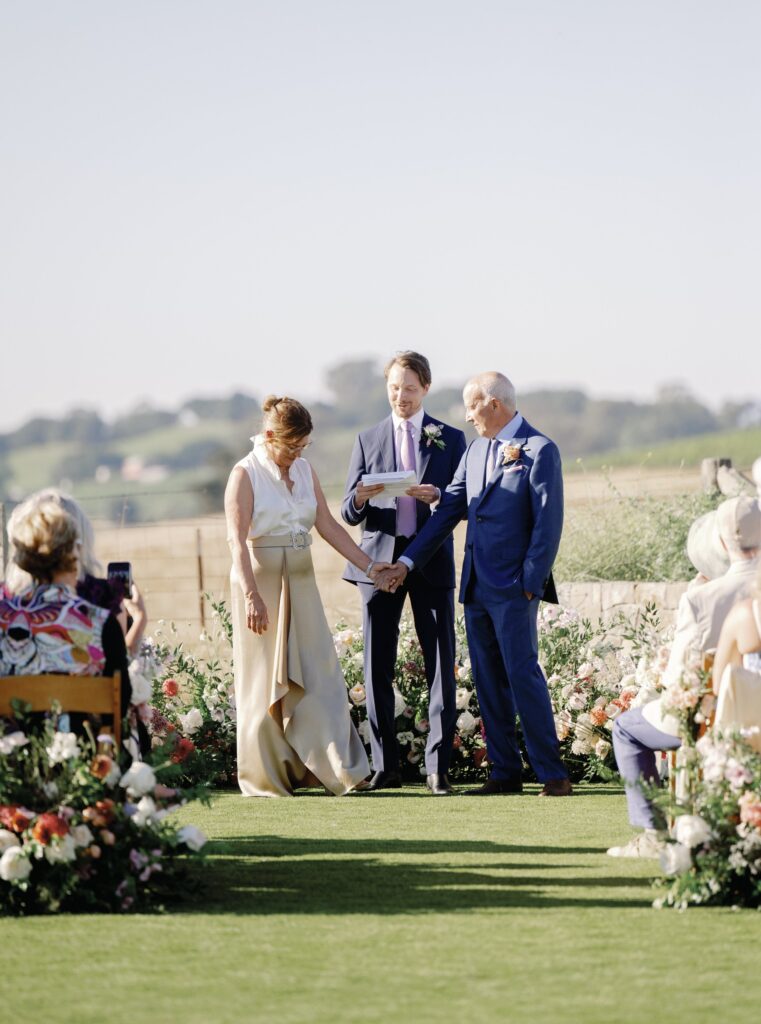 Two people are standing, holding hands, facing each other at an outdoor wedding ceremony. An officiant is present, with flowers and chairs nearby.