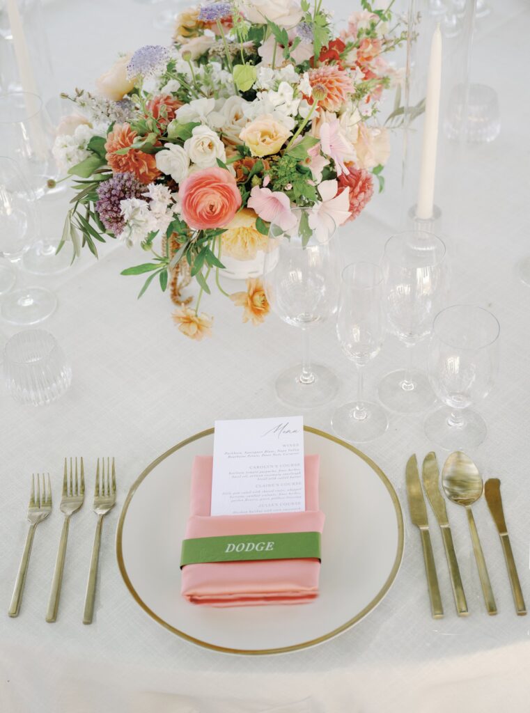 A table setting with a floral centerpiece, menu on a pink napkin, and gold cutlery. The napkin has a green band with the word "Dodge.