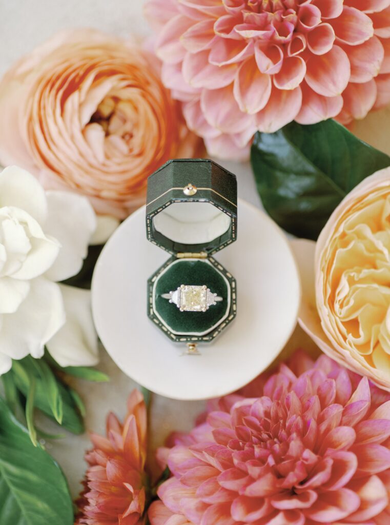 An ornate ring with a square gemstone in an open green box, surrounded by pink and white flowers on a white surface.