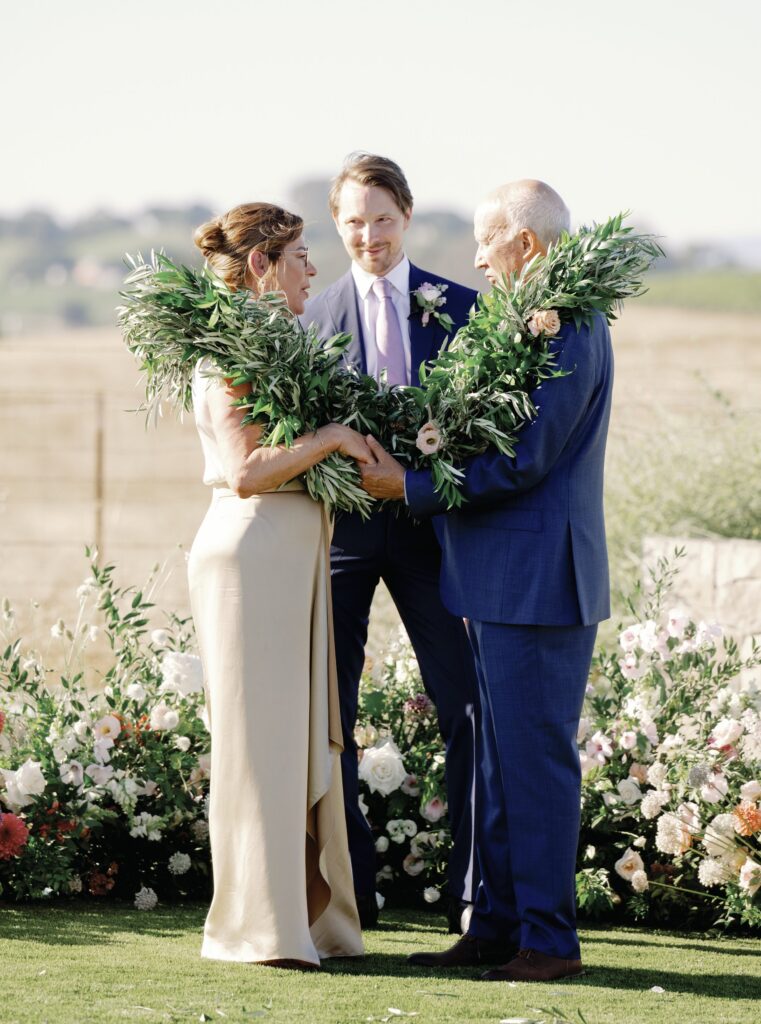A couple stands facing each other outdoors, exchanging large green garlands, with another person standing in the background. There are flowers and greenery around them.