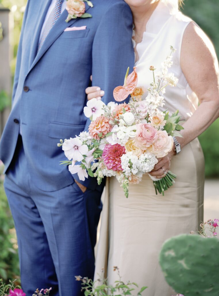 A couple in formal attire embrace, with the woman holding a colorful bouquet of flowers, standing outdoors.