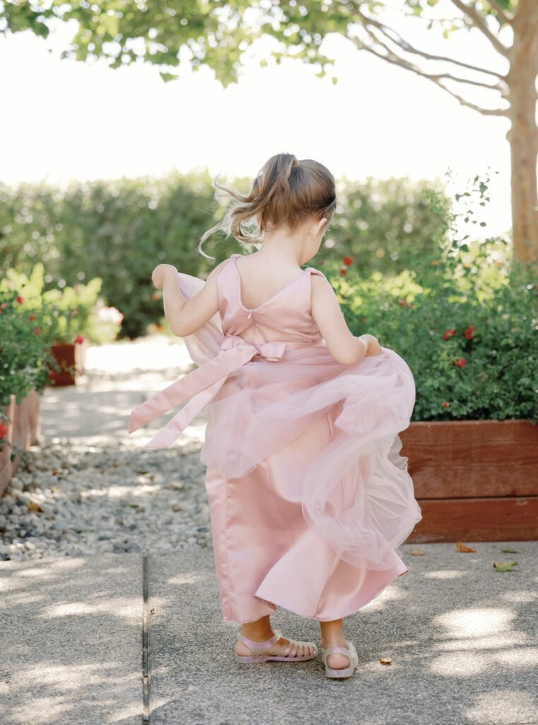 A young girl in a pink dress walks along a garden path surrounded by greenery and sunlight.