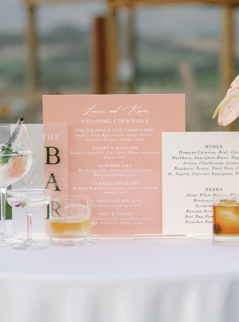 Wedding bar menu on a table with various cocktails, flowers, and a view of a landscape in the background.