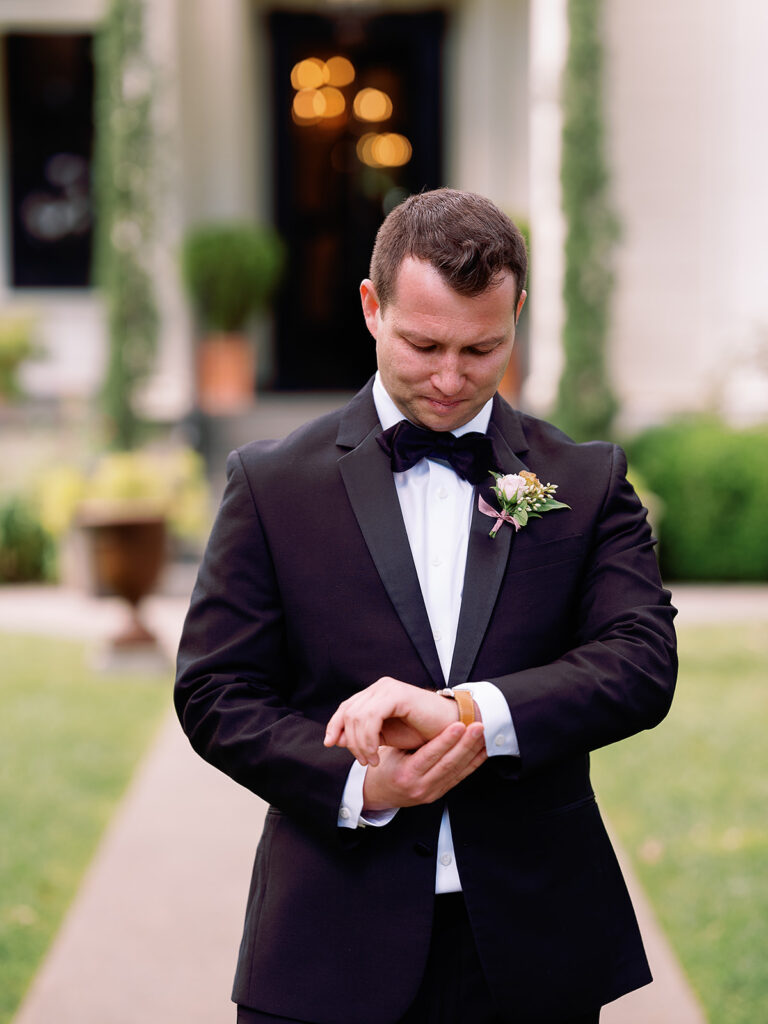 A man in a black suit and bow tie glances at his wristwatch, standing amidst the lush greenery of Park Winters Weddings.