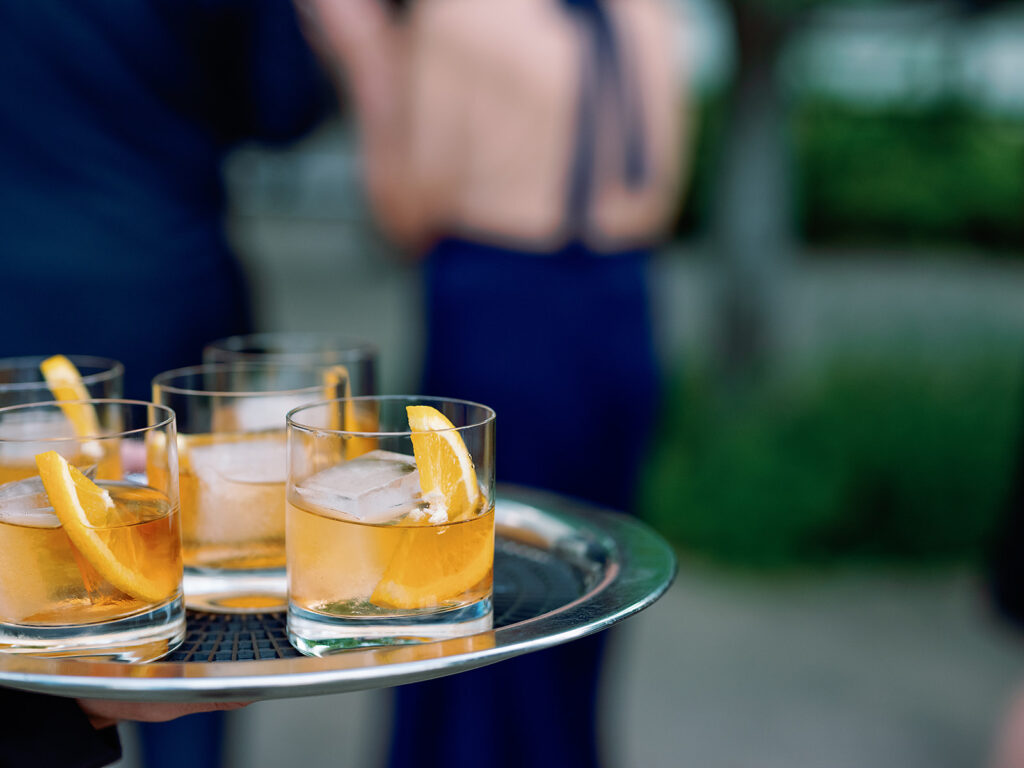A tray with four glasses of iced cocktails, each garnished with an orange slice, is held outdoors at a Park Winters wedding. People are blurred in the background.