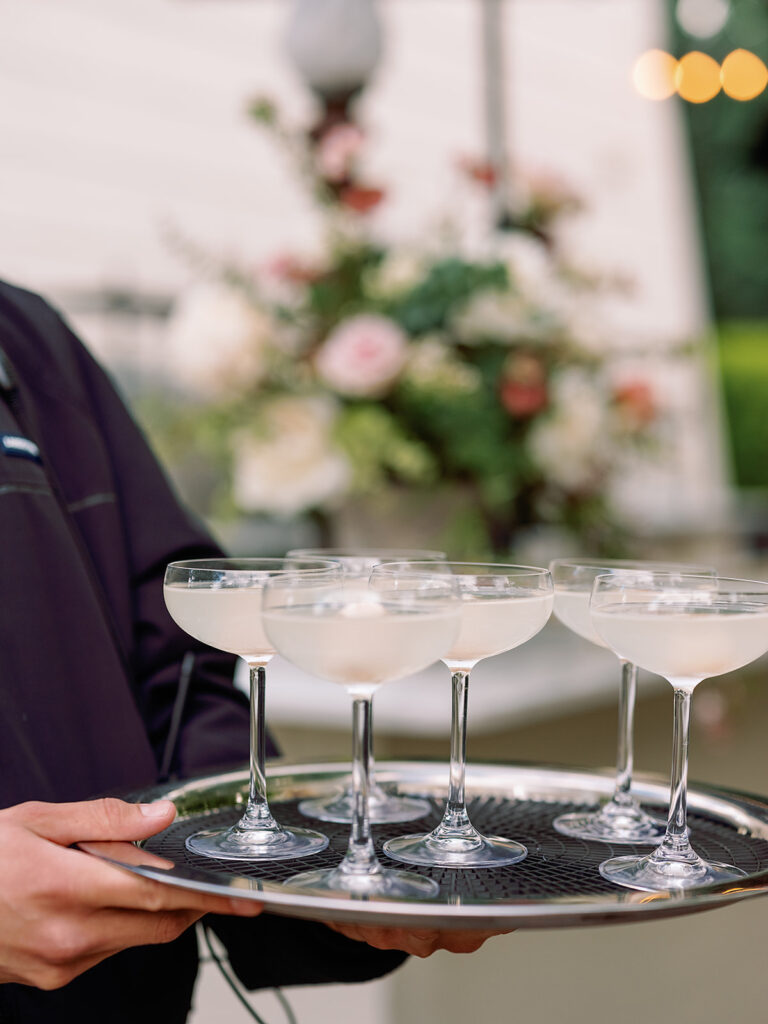 Close-up of a server at Park Winters Weddings, gracefully holding a tray with five champagne-filled glasses, set against a blurred backdrop of flowers and lush greenery.