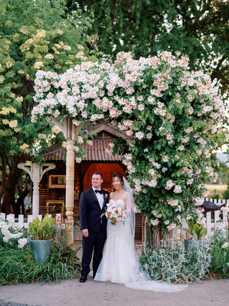 The bride and groom stand under a floral archway at Park Winters, dressed in formal wedding attire, surrounded by lush greenery and a charming picket fence.