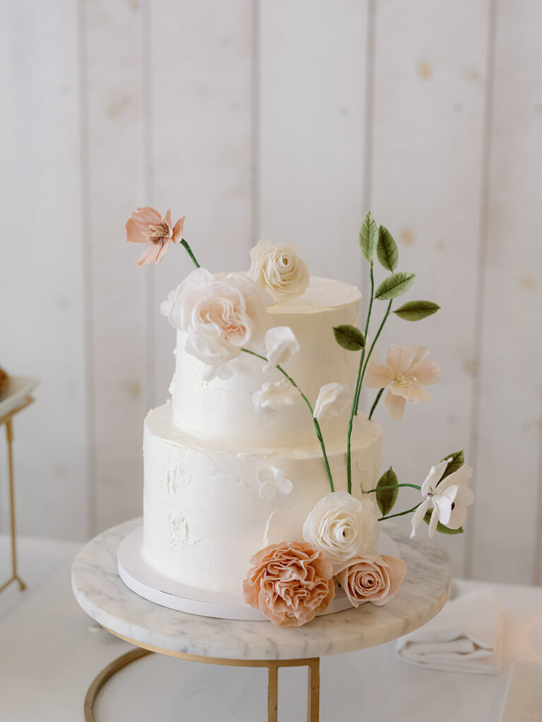 A two-tier white cake adorned with pastel pink and white flowers graces a round marble stand, capturing the enchanting essence of Park Winters Weddings.