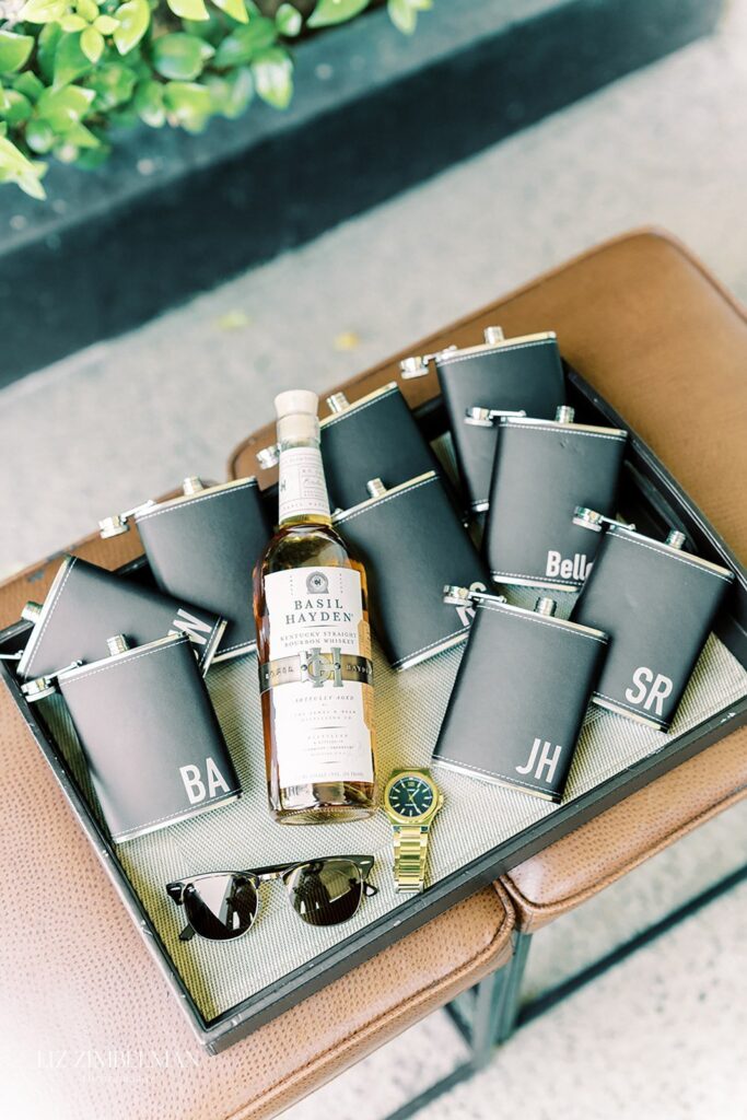 A tray with multiple black flasks, a bottle of Basil Hayden's bourbon, a wristwatch, and sunglasses.