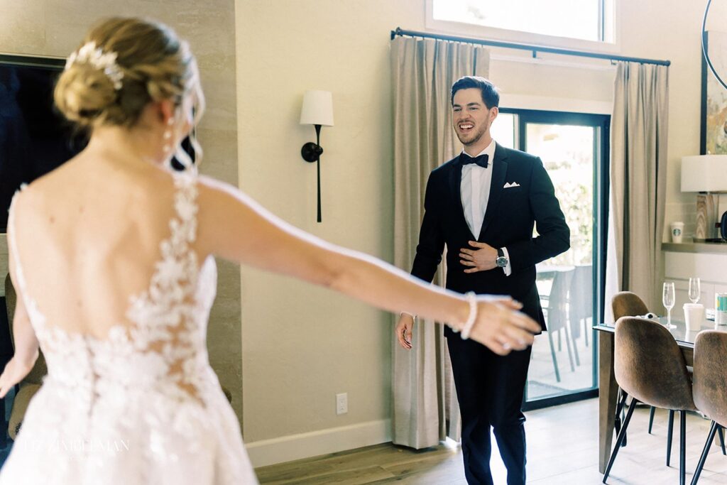 A bride and groom share a happy moment indoors. The bride is in a white dress with lace details, and the groom is in a black tuxedo. They are facing each other with outstretched arms.