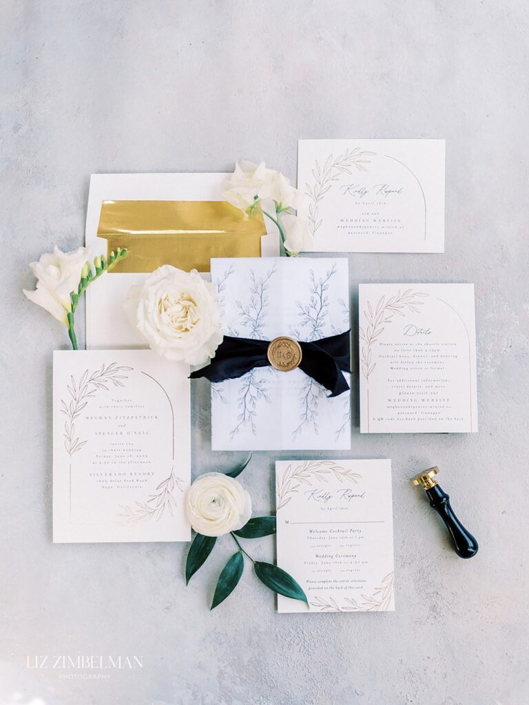 Wedding invitation suite with floral designs, featuring a sealed letter with a black ribbon, surrounded by white flowers and greenery.