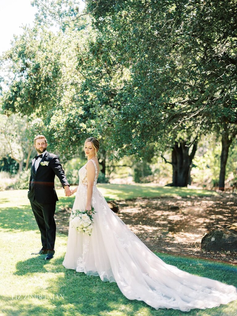 A bride and groom stand hand in hand on a grassy area under trees. The bride wears a long white dress with a train, and the groom is in a black suit.